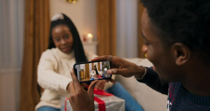 An African-looking guy holds phone and takes photo of girlfriend wrapping Christmas present. The guy took photo and showed it to the girl. A couple in love smiles and rejoices.