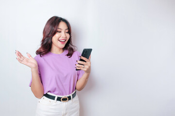 A portrait of a happy Asian woman is smiling and holding her smartphone wearing a lilac purple t-shirt isolated by a white background