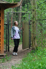 a young beautiful woman in casual clothes and sports sneakers enters the gate and closes it, a green forest remains behind the gate