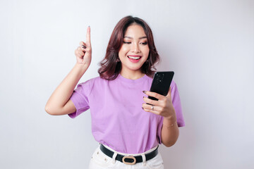 Fototapeta premium Smiling beautiful Asian woman pointing finger upward to empty space while holding her phone isolated white background