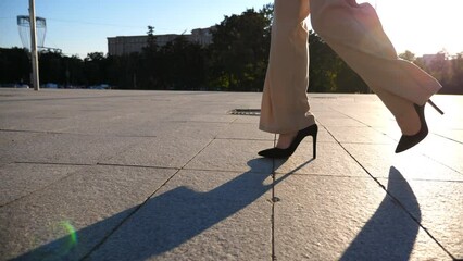 Female legs in high heels shoes walking in the urban street with sun flare at background. Feet of young business woman in high-heeled footwear going in the city. Girl stepping to work. Slow motion