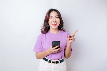 Fototapeta premium Smiling beautiful Asian woman pointing finger to empty space beside her while holding her phone isolated white background