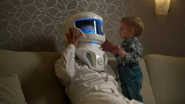 A blond son knocks on the glass of the astronaut's helmet of the father, A man and a boy play astronaut in the interior of a cozy apartment.
