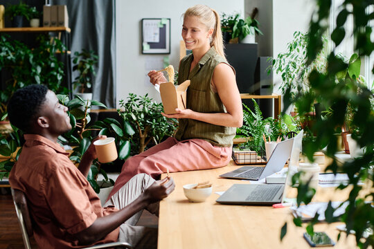 Young smiling female office manager eating wok and talking to African American male colleague sitting by workplace among green plants