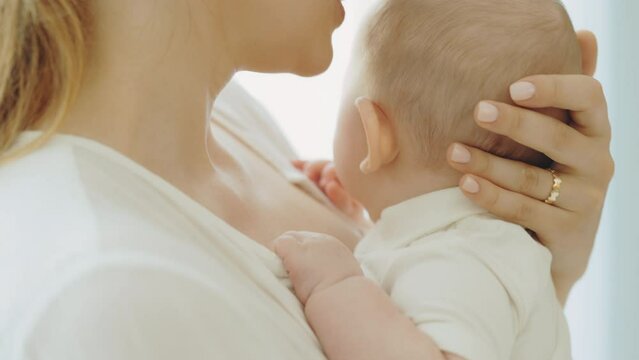 Authentic young mother holding newborn baby in her arms kissing in head, pulls to sleep and care for the child. Close view of the mother holding the baby and kissing her head on the white background