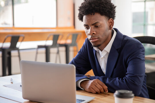 Serious employee sitting at desk and looking at laptop screen. Medium shot of African-American man working in office, thinking, solving problems. Business, technology concept