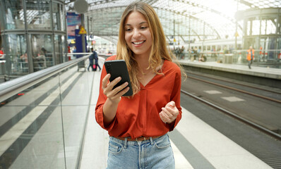 Young smiling woman holding mobile phone on sun flare platform station. Traveler girl on train...