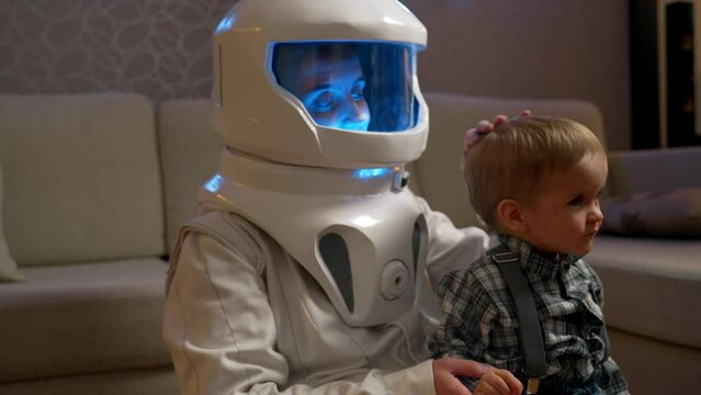 A mother in an astronaut costume pats her son on the head. Playing spaceship at home in the evening. Children's games in adult space professions.