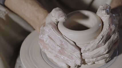 Wet male potter's hands sculpting soft clay and shaping the pot on potters wheel. Master gives shape to the pottery in a creative workshop. Creative masterclass from an artisan. Clay shaping close up.