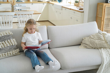 Small girl preschool child reads fairy tale stories holding paper book sitting on sofa alone at home