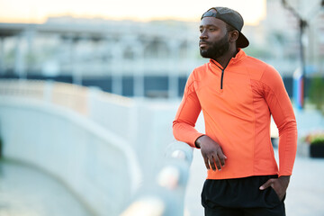 Young serene African American athlete in red sport jacket and black baseball cap standing by riverside and looking at cityscape