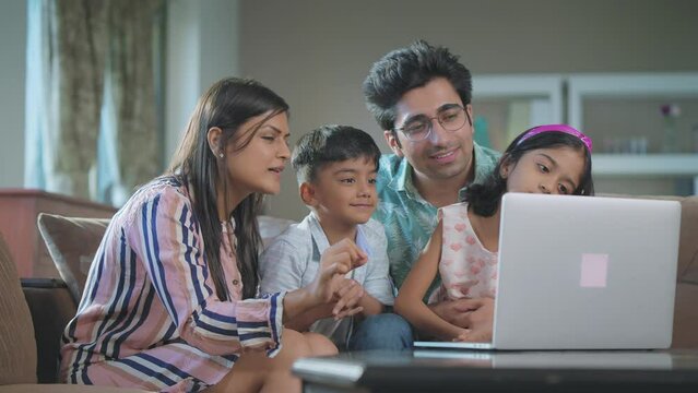 Happy modern young loving Indian Asian family of father, mother, son, and daughter are sitting together on couch watching funny videos or movie on a laptop or notepad in an interior house setup. 