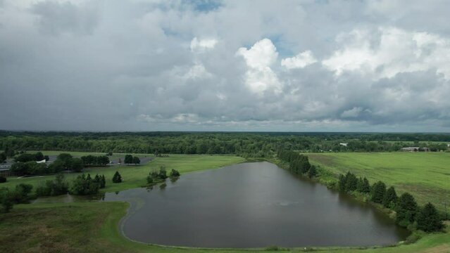 Drone footage shows a still lake close to a busy road that is surrounded by numerous trees, greenery, and an overcast sky.