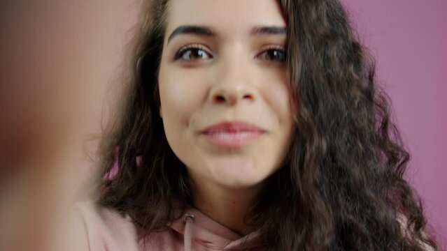 Close-up of excited young lady speaking looking at camera during video call on pink background. People and communication concept.