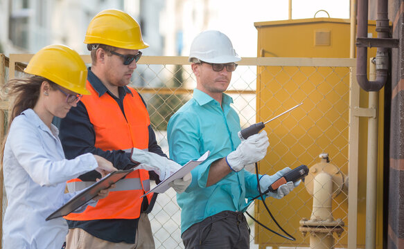 Young Men And A Woman Are Standing Near A Gas Pipe With An Analyzer And Documents. They Are Wearing Helmets, Glasses, Gloves. Checking For Gas Leaks.