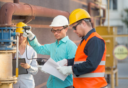 Young Men And A Woman Are Standing Near A Gas Pipe With An Analyzer And Documents. They Are Wearing Helmets, Glasses, Gloves. Checking For Gas Leaks..