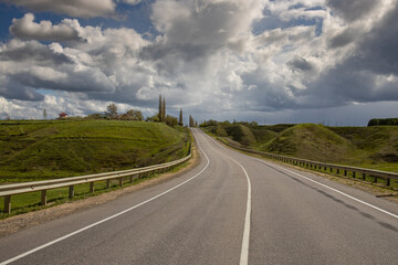Osphalt road. Spring landscape, dramatic sky over green hills, before rain. The sun through the clouds illuminates the road and ravines.