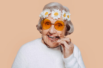Closeup portrait of cheerful senior old grandmother bites a metalic symbol of peace necklace. Hippie elder woman with wreath on her head. Funny portrait.