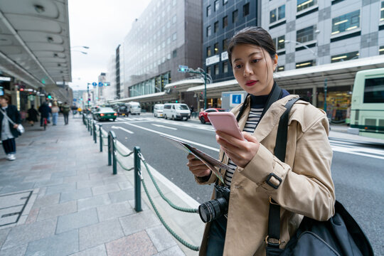 asian Japanese woman traveler holding map and checking departure time on her cellphone at a bus station near the street in downtown Kyoto city in japan