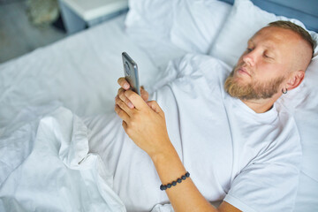 Bearded man using smartphone in morning in cozy while lying on bed