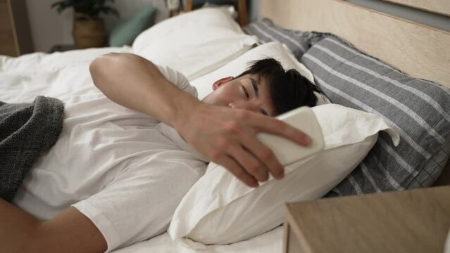 sleepy asian young guy woken by alarm clock triggered on his smartphone keeps sleeping after checking time and switching it off in the bedroom at home