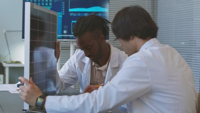 Two diverse male electrical engineers in lab coats having discussion while examining solar panel and technical drawings in office, working together on sustainability project