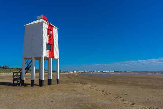 Standing on stilts, the wooden lighthouse on the beach near Burnham on Sea. The lighthouse is painted white with a single vertical red stripe on its front. You can see the seafront at Burnham in the d