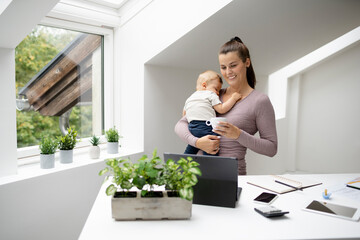 pretty young woman, businesswoman stands at work desk holding her sleeping son in her arms and working on notebook, smartphone and is happy