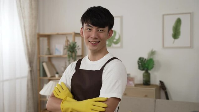 half length portrait of a professional asian male housekeeper in apron and gloves is posing with folded arms and smiling at camera at customer’s house.