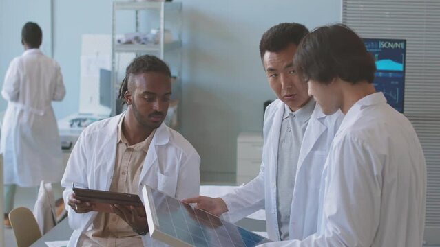 Team of multiethnic electrical engineers in lab coats examining solar panel while developing environmental project together in office
