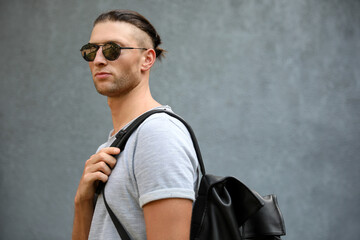 Handsome young man in stylish sunglasses with backpack near grey wall, space for text