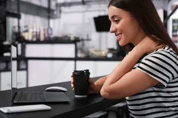 Happy young woman with paper cup of coffee and laptop at table in hostel dining room