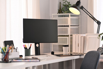 A blank computer screen, stationery and lamp on white table in modern home office interior