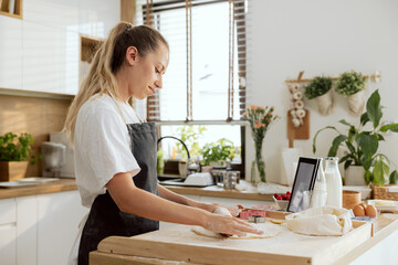 Happy woman in the kitchen wearing apron rolling out dough preparing for homemade cookies, cakes, pizza. Preparing lunch for kids. Looking at tablet with recipe.