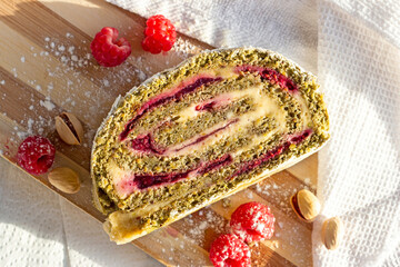 Green pistachio meringue roulade slice with raspberry jelly jam on light wooden board background in...