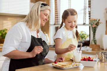 Eldest grandmother and happy grandchild baking cookies in the kitchen, pouring milk in glass, preparing breakfast for mother. Women having fun.