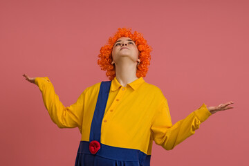 a girl in a clown costume and a bright wig looks up and spread her arms to the sides, studio portrait on a colored background