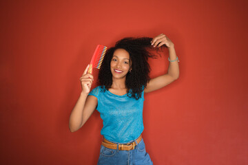 Young happy black american woman combing her curly afro hair on red background. Charming african...