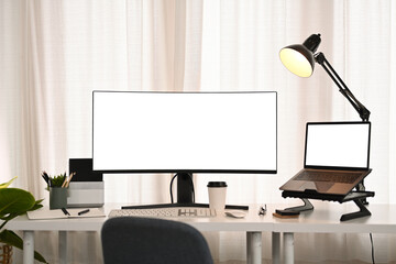 Ultra wide monitor, laptop, stationery and lamp on white table in modern creative office
