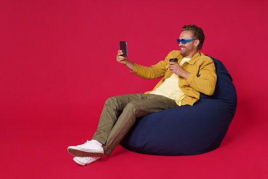 Happy handsome young man 30s make selfie using digital tablet drinking coffee from paper cup wearing casual denim outfit sitting on bag chair isolated on red background. Coffee shop business concept