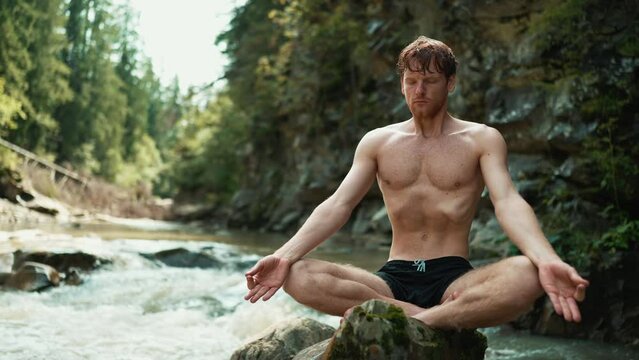Pretty red haired man doing yoga meditation on a stone in the running river 