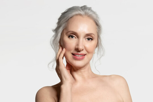 Gray-haired beautiful senior female touching perfect skin. Smiling mature good-looking lady enjoying groomed face. Anti-wrinkle care, cosmetics for elderly. Close up studio portrait on white