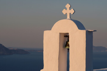 A cross and a bell on the top of an orthodox church in Ios Greece while the sun is setting in the background - obrazy, fototapety, plakaty