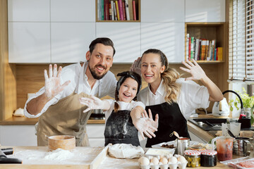 Delighted father and mother teaching curious daughter cooking baking. Happy family in aprons kneading homemade dough for pizza pasta gnocchi. Kitchen tools sieve on wooden surface.