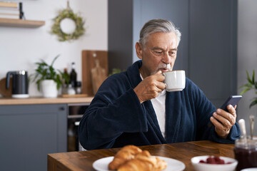 Senior caucasian man eating breakfast at home and using mobile phone