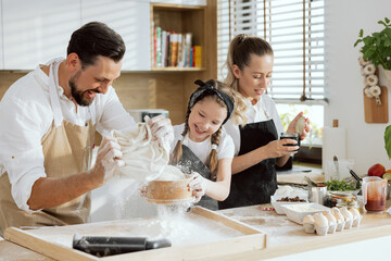 Family having fun together. Daughter and happy father sieving flour on wooden surface. Blonde mother opening jam. Prepared ingredients on wooden table. Modern light kitchen with large window.