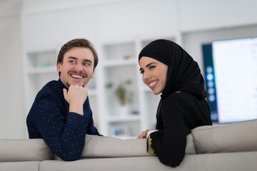 Young muslim couple woman wearing islamic hijab clothes sitting on sofa watching TV together during the month of Ramadan at modern home