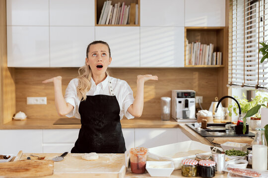 Confused young lady woman chef with opened- mouthed holding hands near head looking at camera in amazement wearing apron cooking baking homemade pizza cookies.