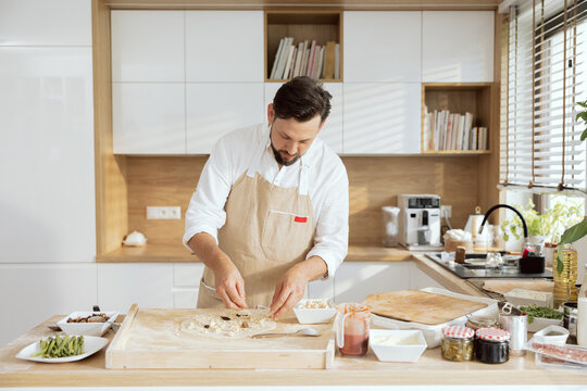 Handsome chef with beared dressing pizza with feta cheese mushrooms olives salami preparing homemade pizza in modern kitchen at home. Having fun relaxing alone. Wearing apron.