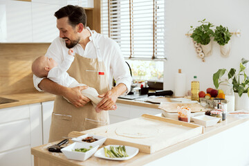 Happy father in aprons looking at adorable baby holding in arms standing at wooden table cooking baking homemade pizza with mushrooms salami and tomato sauce.